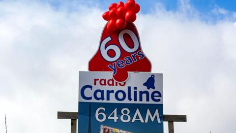 Radio Caroline billboard celebrating the station's 60th birthday in 2024, it is blue and white with red balloons at the top.