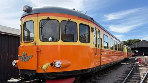 A orange rail cart, made up of two carriages, is on the tracks on a sunny day. It is rounded at the sides with yellow paint around the windows, orange on the body and black on the top and bottom. 