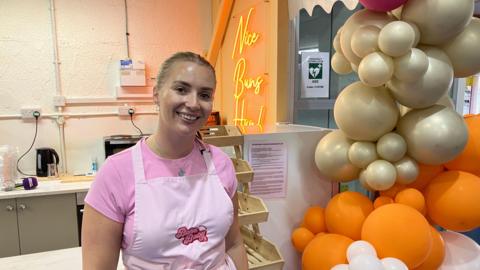 A woman in a pink t-shirt and apron smiles at the camera, in front of a small kitchen in a marketplace. A large display of gold, orange and light pink balloons are next to her.