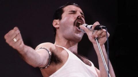 Freddie Mercury sings into a microphone, his eyes shut tightly and his right hand in a fist, held out to the side. He is wearing a white vest and has a studded armband around the upper half of his arm.