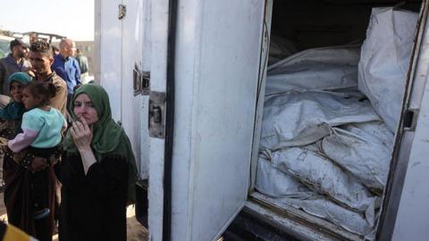 A Palestinian woman stands next to a refrigerated lorry carrying the remains of unidentified Palestinians whose bodies were returned by Israel in exchange for dead Israeli hostages, outside Naser hospital, in Khan Younis, southern Gaza (22 October 2025)