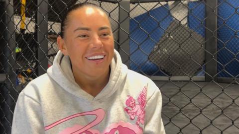 Kennedy Freeman, wearing a cream hoodie with a pink logo, standing in from of wire fencing, smiling.