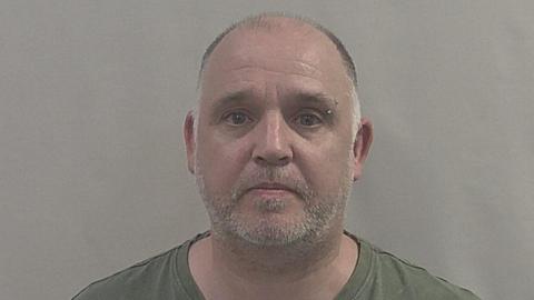 A police headshot of Stacie John Farrar. He has short/ balding dark-coloured hair on his head and short grey facial hair. He is wearing a khaki green t-shirt. 