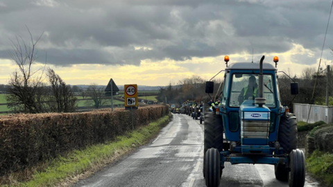 A line of tractors driving down a country road beneath a cloudy sky. Houses are visible on the right of the image while a hedge and fields can be seen on the left. 