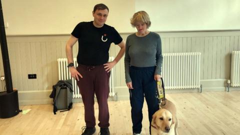 Darren Murphy (left) and Andrea Hope (right) are standing in their dance studio on a wooden floor with panelled walls and radiators behind them. Mr Murphy has short brown hair and Ms Hope has short blonde hair. She is holding the harness of a golden Labrador guide dog. A grey backpack rests on the floor to the left near the wall behind them.