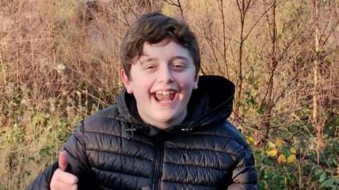 Kyle sitting on a rock overlooking his hometown, Pontypridd. He has short black hair and is wearing a black puffer coat. He is smiling and making a thumbs up gesture.