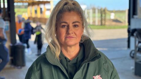 Heather Hogarty has her straight blonde hair in a ponytail as she looks at the camera with a serious expression. She is wearing a khaki hoodie and jacket. Farm visitors can be seen behind her.