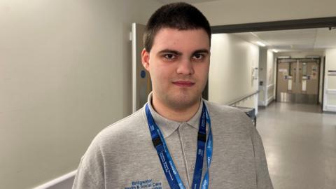 A man wearing a grey polo T-shirt with two blue lanyards is standing in the corridor of a hospital. He has dark brown hair, dark eyes and is smiling at the camera.