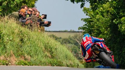 Dean Harrison in red and blue leathers riding away on the TT course. He is leaning over towards a grass embankment where a group of spectators are holding up mobile phones filming him.
