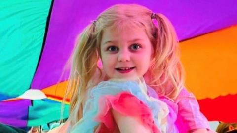 A little girl with blonde pigtails. Dressed in a tutu, she is sitting under a colourful parachute, looking at the camera and smiling. 