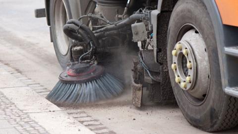 A road sweeper at work. A rotating brush attached to the side of the vehicle is cleaning the gutter of a road.