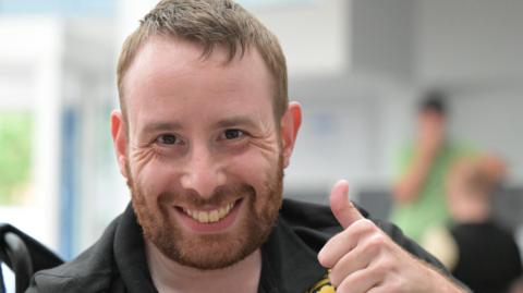 A man with short light brown hair is giving a thumbs-up gesture while seated indoors. He is wearing a black polo shirt with gold embroidered logos, including one circular badge on the chest. The background is softly blurred, showing another person in green.