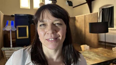 A woman with dark shoulder-length hair is facing the camera with her exhibition in the background. It is a head and shoulders shot of her. She has a cream shirt on. 