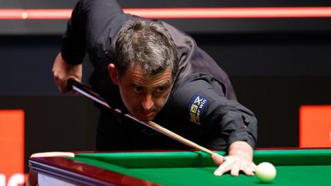 Snooker player Ronnie O'Sullivan prepares to strike the white ball. He has black hair, which is greying in parts, stubble, and is wearing a black shirt and waistcoat. 