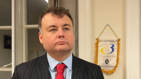 A man with dark hair, a dark suit and a red tie standing in a room with white walls and a small pennant hung behind him