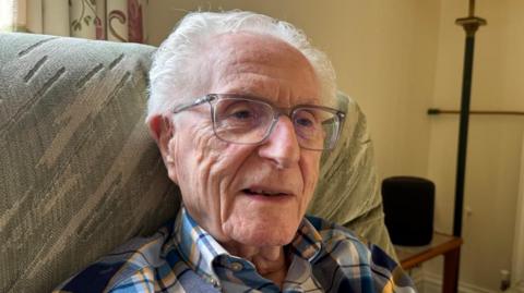 A man sitting in a chair wearing glasses and a check shirt, looking slightly to the side of the camera.