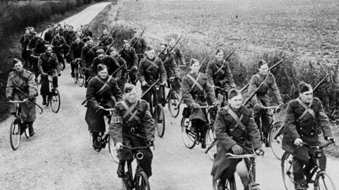 A black and white image of a group of men in wartime Army uniforms, with rifles slung over their shoulders. The are all cycling up a line with a hedgerow and field on the right, near Sudbury in Suffolk, in 1941.