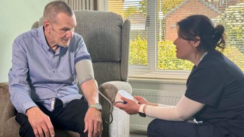 The doctor is sat in the front room with David Clayton testing his blood pressure