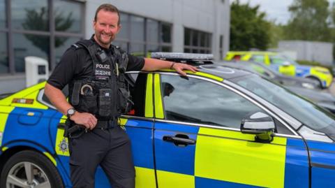 PC Paddy Connell in uniform. He is standing beside a police patrol car and is smiling. There is a grey building and another police vehicle in the background. PC Connell has brown hair and a short beard.