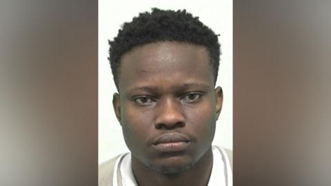 Mugshot if Ishiekwenne. He has very faint black stubble on his chin and upper lip and short thick black hair.
