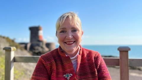 Tricia Warwick is sitting in the sunshine in front of Archirondel beach, where there is a fence, a tower and the sea in the background. She is wearing a red jacket and red lipstick and is smiling at the camera.