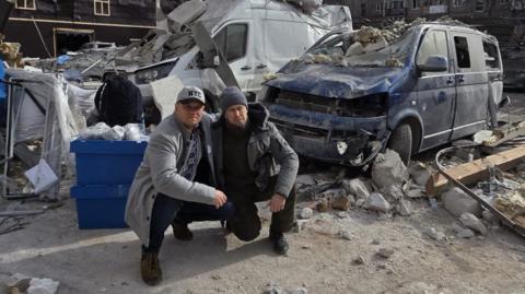 Karol Swiacki and March Edwards crouching in front of their destroyed vehicles