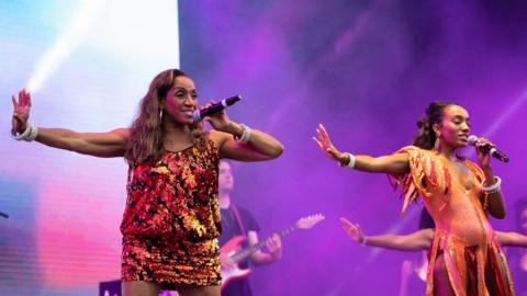 Sister Sledge dancing on stage wearing orange outfits. There is a purple background behind them.