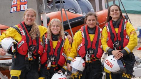 RNLI crew members Anna Heslop-Latif, Sarah Whitelaw, Karen Pearce and Jane Heslop-Latif. The women are wearing yellow and black uniforms and red lifejackets. They stand in front of a RIB outside the Cullercoats station.