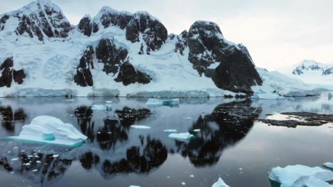 The Antarctic ocean. There is some ice in the water. In the background there is land covered in snow and ice