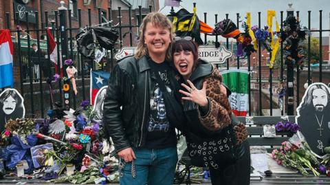 A couple at the Black Sabbath Bridge. The man has long blonde hair and wears a black leather jacket. He has just proposed. The woman has dark hair and a leopard print coat. We can see the Black Sabbath Bridge behind them. She has holding her hand up. 