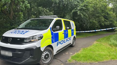 Police van in park with blue and white police tape behind it