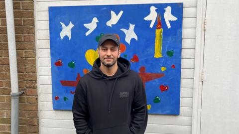 Tarik Alali in a baseball cap and black hoodie standing outside in front of a blue painting with doves and a candle.