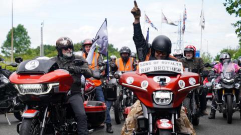 Hairy Biker Si King on his motorbike at the National Motorcycle Museum in Solihull during Dave Day. Next to him is another rider who is raising his arm and pointing his index finger in the air. Behind them are other bikers.