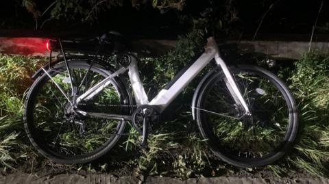 A modified e-bike lies on the ground, on grass on the hard shoulder of a motorway at night time. The wheels are black and the frame is silver in colour. The back break light is glowing red across the floor. 