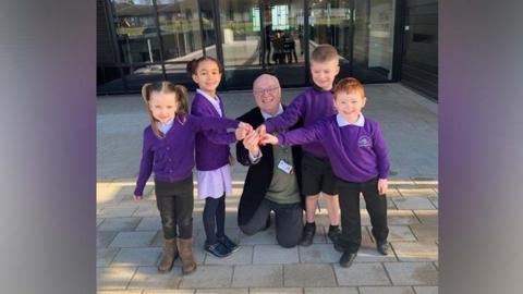 Four children in purple jumpers and a man in a black blazer all hold a key infront of a building with glass doors and windows behind. 