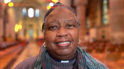 Rev Eileen Cremin is smiling at the camera. Her hair is tied back and she is wearing a grey coloured jumper. 