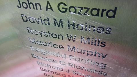 Black names engraved onto a silver metal plaque with scratches all over them, as if an attempt has been made to scratch them out.