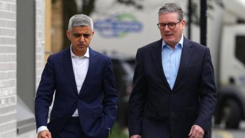 Sir Sadiq Khan and Sir Keir Starmer walking side-by-side on a public street. A lorry is seen out of focus in the background and they are next to a grey bricked building.