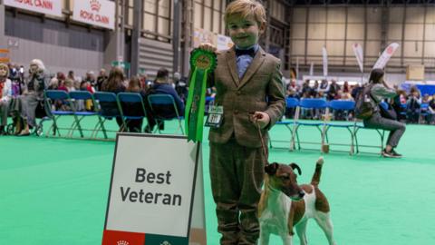 A young boy in a tweed suit called Freddie holds a green rosette and the lead of his smooth haired fox terrier Penny. He stands on a green carpet at a dog show next to a sign reading "Best Veteran". 