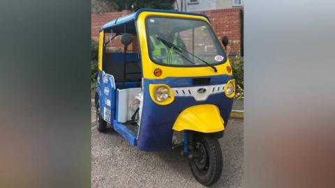 A blue and yellow painted tuk-tuk is parked on a road. A police officer is inside, wearing a high visibility jacket. Behind him are plants and a brick wall.