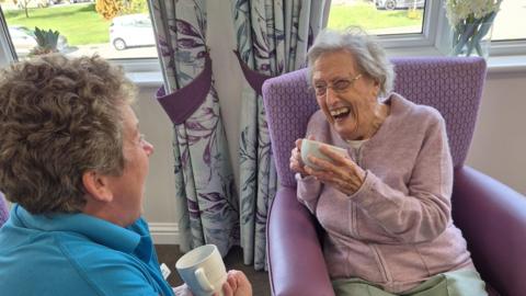 A woman with grey hair and glasses with a big open smile, wearing a lavender zip up top and light green trousers as she sits in a purple chair. She is looking at another lady with cropped wavy hair who has her side to the camera. 

The woman is wearing a blue top. They are both holding mugs in their hands as they seem to be enjoying a joke together. 
In the background, there are a series of windows and curtains with purple leaves and purple curtain ties holding them open. There is also a glass vase with a bunch of flowers on the window sill.