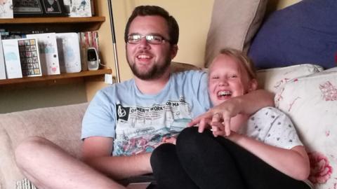 A man with short brown hair, stubble and rectangular glasses is wearing a blue top and smiling on a sofa with his arm around a teenager. The teenager has her mouth open laughing with braces and has blonde hair tied back, a white top and black leggings. Their hands are entwined. Next to them is a shelf with cards and books on it.