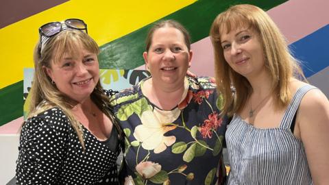 A blonde woman in a black and white dotted shirt, is standing next to a blonde woman in a flowery print shirt. On her right is another blonde woman wearing a striped white and dark navy top. 