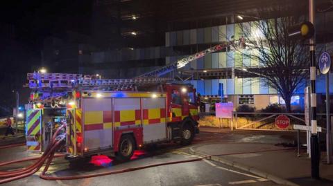 A fire engine with hoses running from the back is parked facing a car park building with a ladder extended towards it from its roof.