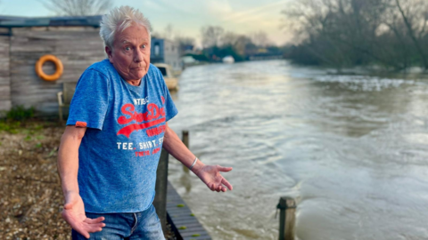 Andy Ellison, standing next to the River Thames, where the boat was once moored, has his hands out as if questioning where the boat has gone