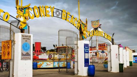 Entrance gates to amusement park with Coney Beach Pleasure Park in yellow letters on top of the gates and attractions visible behind 