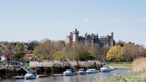 Several boats moored on a river. There is a stone castle in the background.