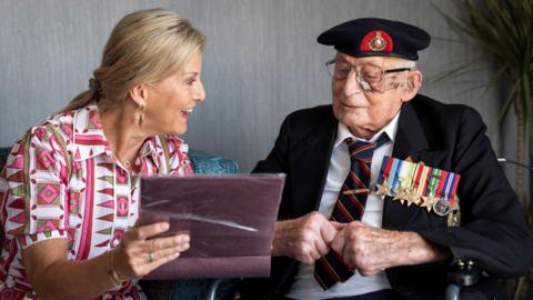 The Duchess of Edinburgh sits next to 105-year-old Royal Marines veteran James 'Jim' Wren. She is holding a photo album and going through it with him, She wears a dress patterned with pink and he is in uniform with multiple medals.