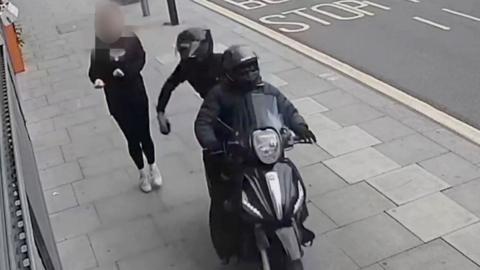 A CCTV image showing a black moped being driven along a pavement by two people wearing all black, snatching a mobile phone from a pedestrian walking along the pavement. One of the people on the moped can be seen holding the phone in their hand moments after snatching it, as the pedestrian recoils.