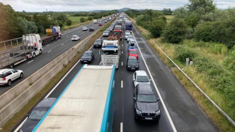 Three lanes of a motorway are jammed with black, white, red and blue cars and lorries. The queues stretch into the distance. Traffic can be seen travelling normally in the opposite three lanes on the left. Green fields and bushes line the motorway.